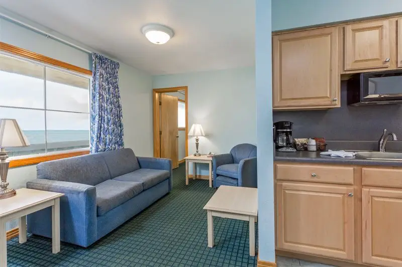 A suite living room at Mariner Inn & Suites with a blue sofa, an ocean-view window with a patterned curtain, and a kitchenette with a coffee maker
