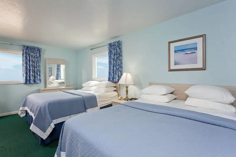 Two beds with pale blue coverlets at Mariner Inn & Suites, with a framed print of a small boat on a beach above the headboard