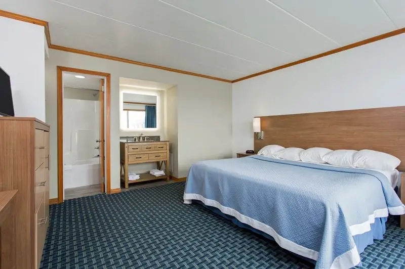 A king bed with a blue coverlet at Mariner Inn & Suites, with the bathroom visible through an open door and a wood vanity nook alongside