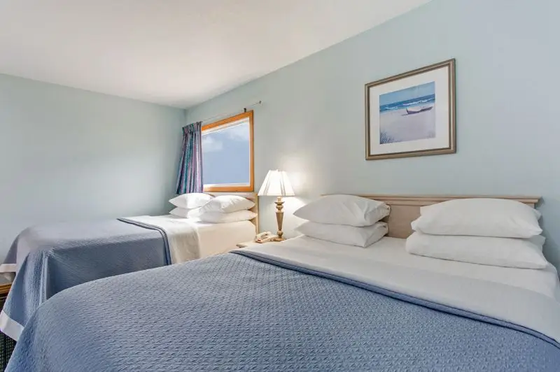 Two beds with blue coverlets at Mariner Inn & Suites, with a framed beach print above the headboard and a table lamp between the beds