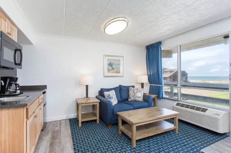 A suite at Mariner Inn & Suites with a teal armchair and sofa facing an ocean-view window, and a full kitchen with a refrigerator and microwave to the right