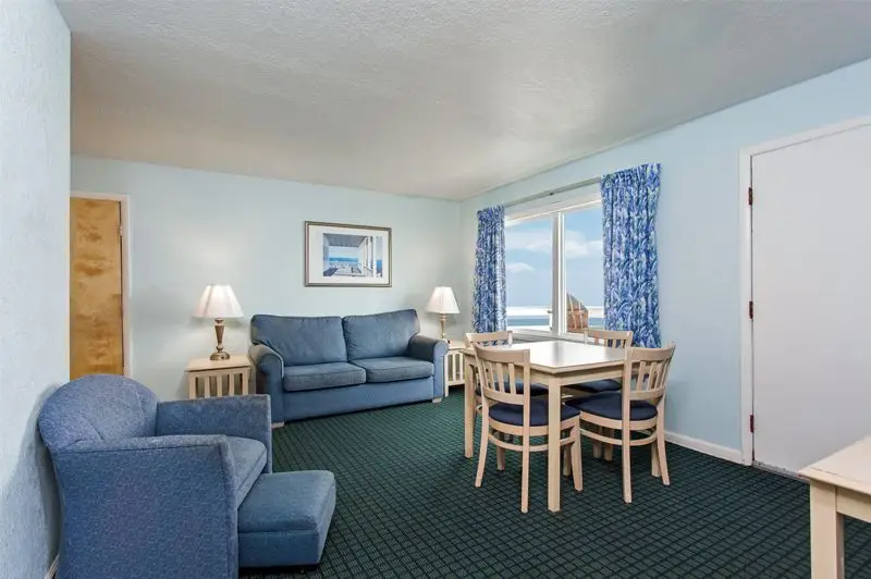 A suite living room at Mariner Inn & Suites with a blue sofa and armchair, a dining table for four, and an ocean-view window with a floral curtain