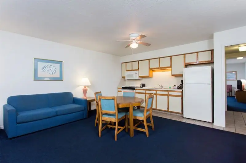A suite living and dining area at Mariner Inn & Suites with a navy sofa, a wood dining table, and a kitchen with a white refrigerator visible through the doorway