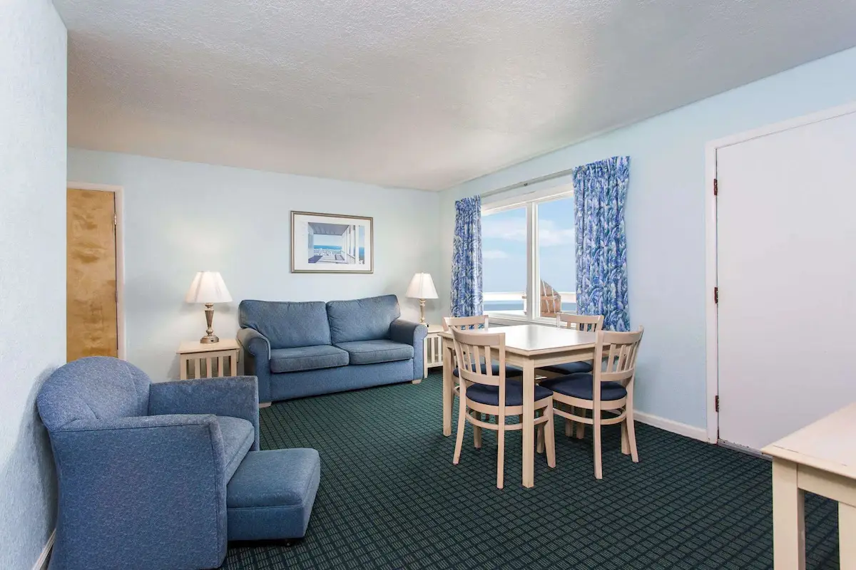 A suite living room at Mariner Inn & Suites with a blue sofa and armchair, a dining table for four, and a floor-length window with a floral curtain