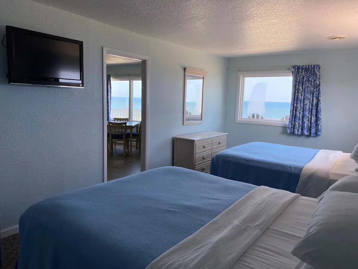 A bedroom at Mariner Inn & Suites with a wall-mounted TV and dresser, opening onto a living and dining area with an ocean-view window
