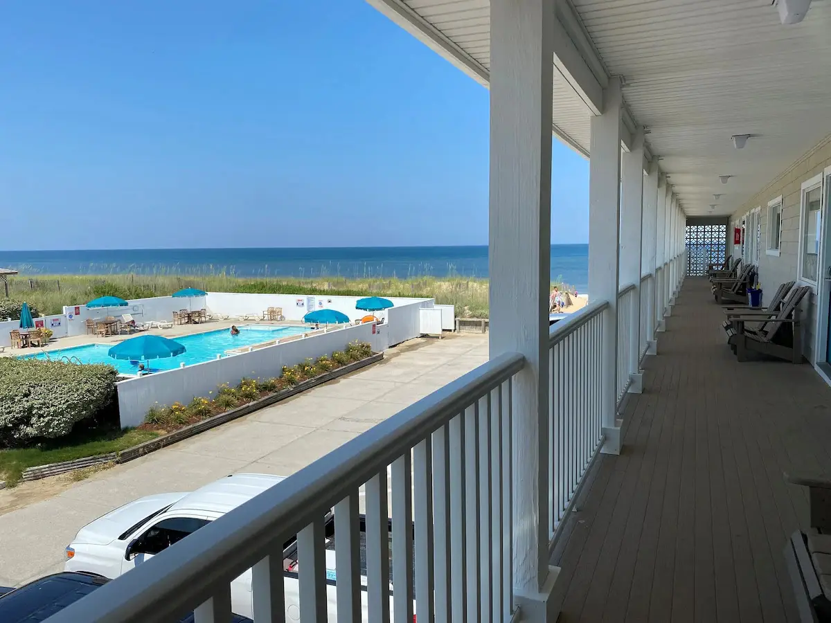 The covered upper-floor walkway at Mariner Inn & Suites, lined with wooden porch chairs outside guest room doors, overlooking the pool and the Atlantic Ocean