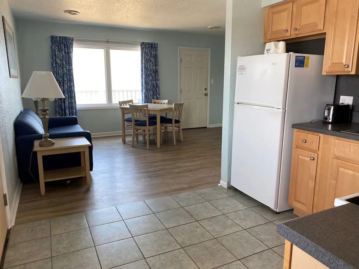 A suite living and dining area at Mariner Inn & Suites with a navy loveseat, a dining table, and a floor-length ocean-view window with a floral curtain