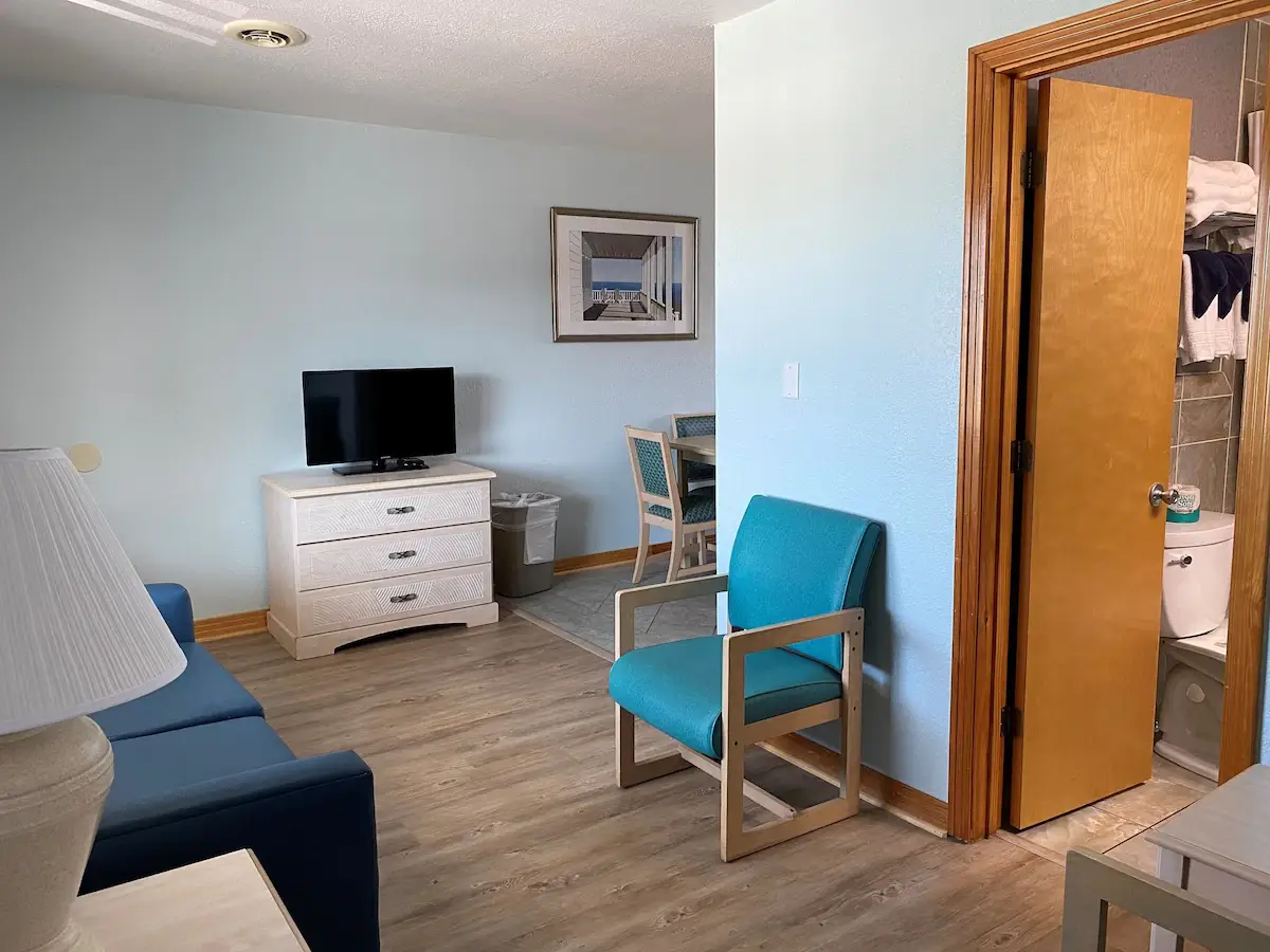 A suite living room at Mariner Inn & Suites with a TV on a white dresser, a teal accent chair, and the bathroom door open at right showing towels