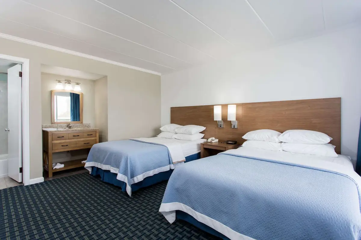 Two beds with blue coverlets at Mariner Inn & Suites, with a wood vanity and mirror nook and dark patterned carpet