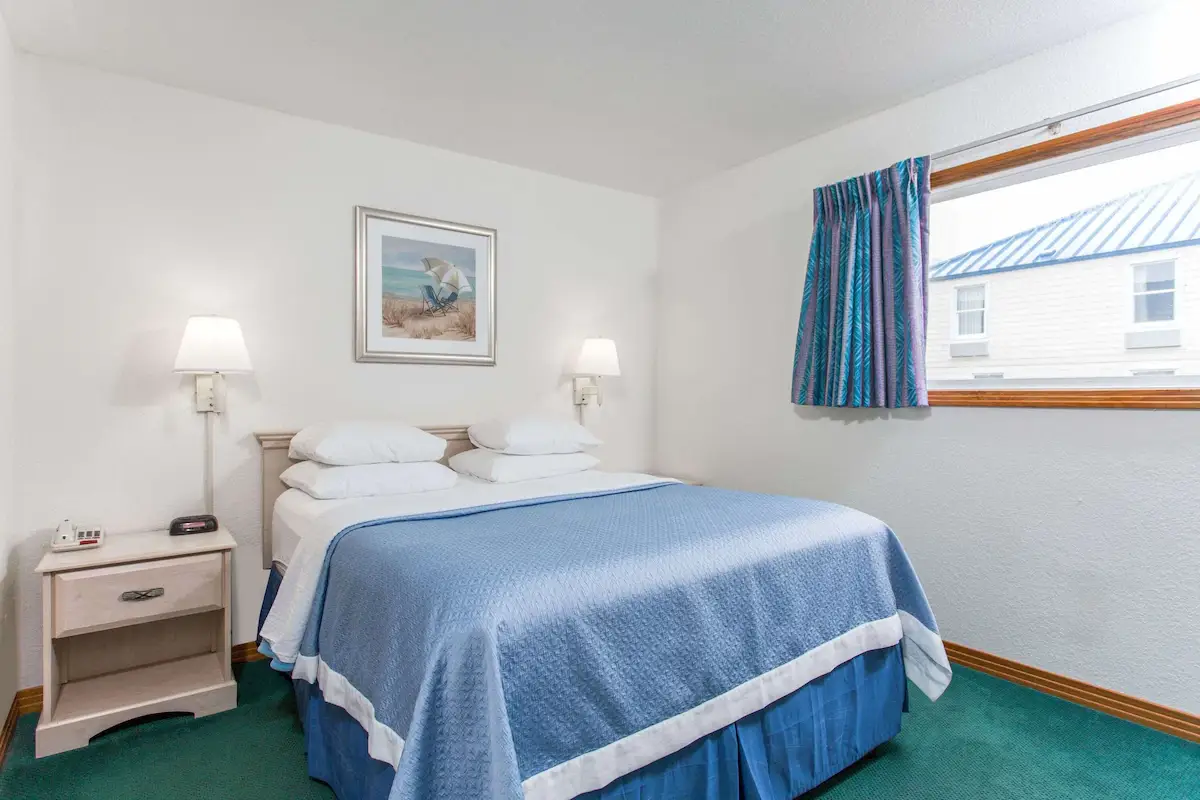 A king bed with a pale blue coverlet at Mariner Inn & Suites, with a framed beach-chair print above the headboard and a window showing a neighbouring rooftop