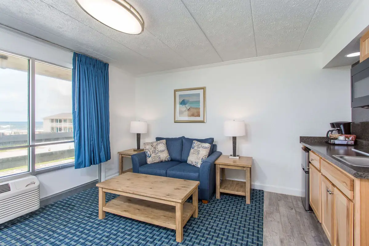 A suite living room at Mariner Inn & Suites with a blue loveseat, a teal accent chair, a small round table and an ocean-view window