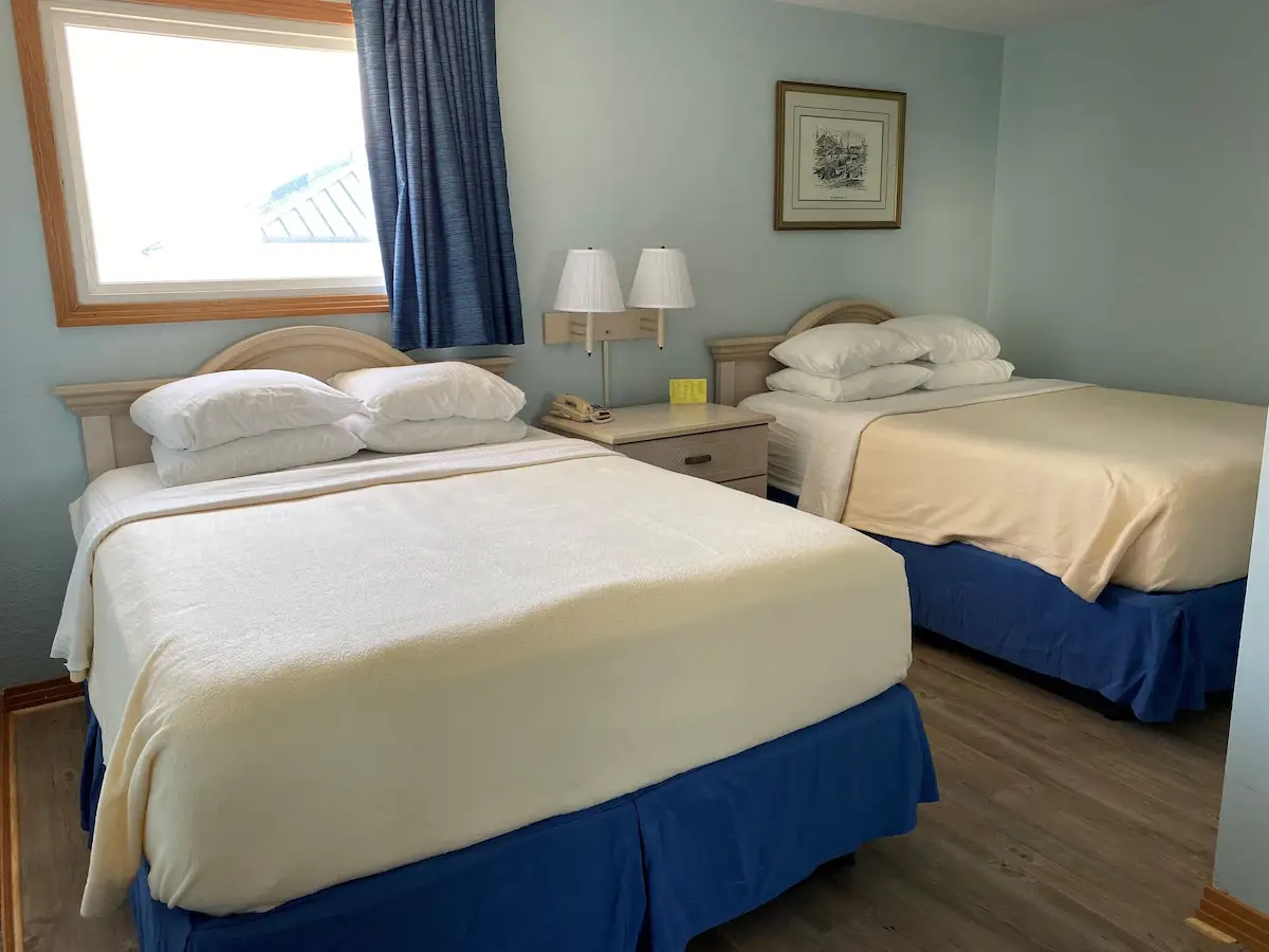 Two beds with pale coverlets and blue skirts at Mariner Inn & Suites, with a black-and-white framed print above the headboard