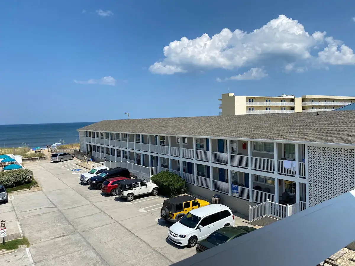 An elevated view of Mariner Inn & Suites, a two-storey building with blue shutters and balconies, the ocean to the left and a parking lot full of cars
