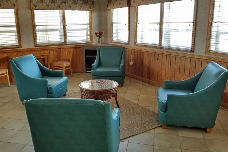 A guest sitting room at Mariner Inn & Suites with four teal wingback chairs around a round wood table, wood-panelled walls and large windows