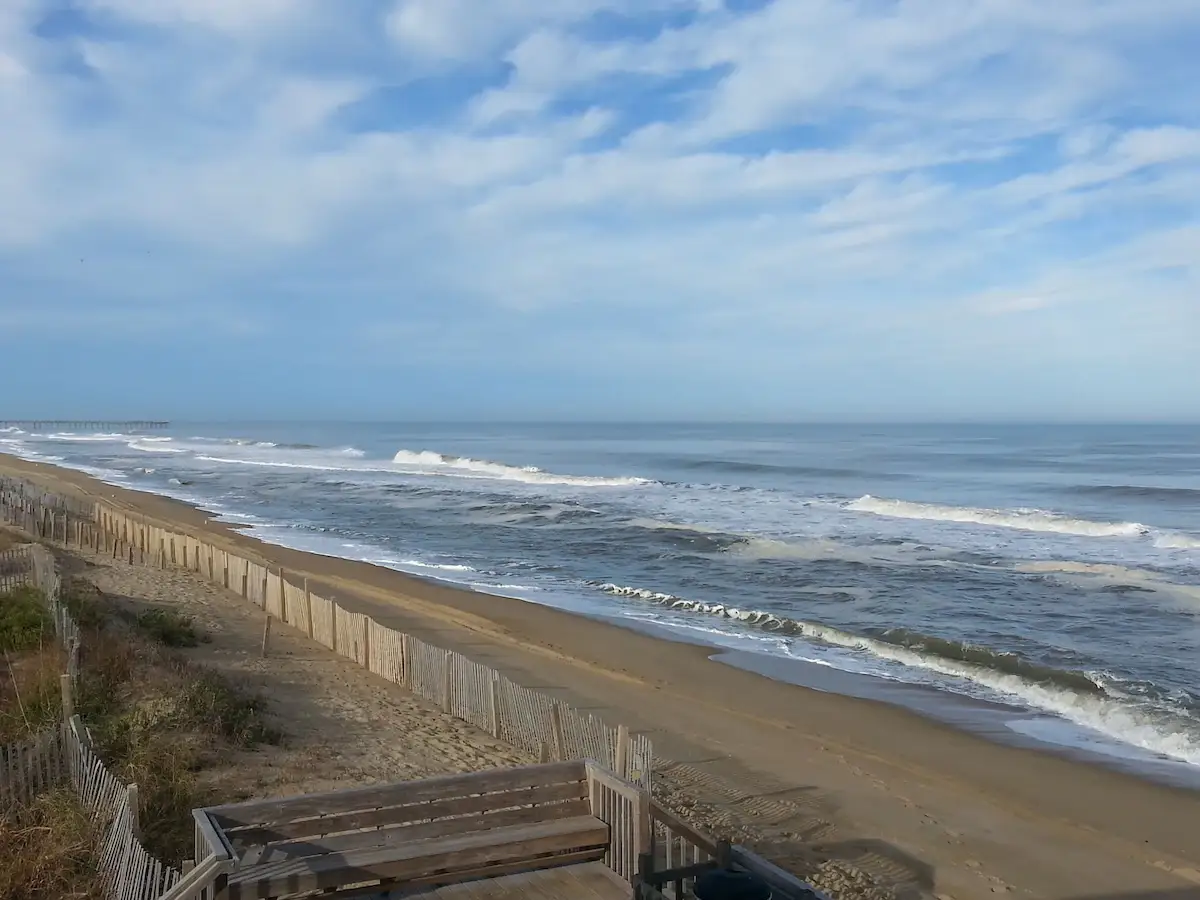 The beach in front of Mariner Inn & Suites, with a wooden dune fence in the foreground, waves rolling in, and a fishing pier visible in the distance