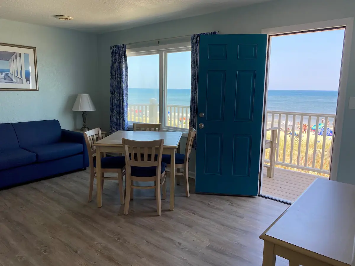 A suite living room at Mariner Inn & Suites with a navy sofa and dining table, and the teal front door open to a private balcony overlooking the beach and the Atlantic Ocean