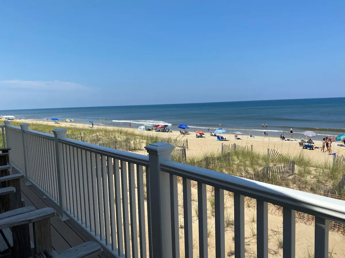 The view from the hotel's own boardwalk at Mariner Inn & Suites — wooden steps and a white railing leading down past the dune fence and sea oats to a busy beach of coloured umbrellas, and the Atlantic beyond