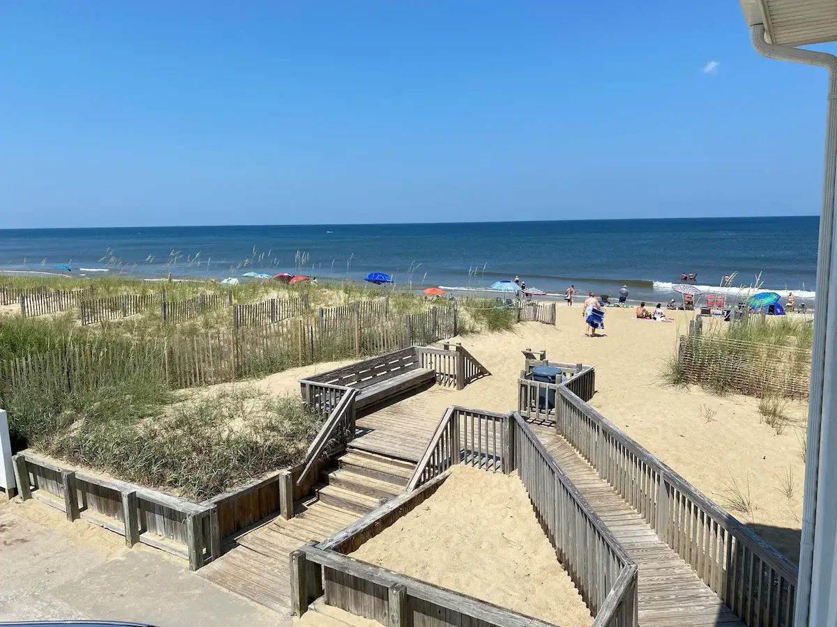 A wooden boardwalk and steps leading down through dune grass to the beach at Mariner Inn & Suites, with beachgoers and umbrellas in view