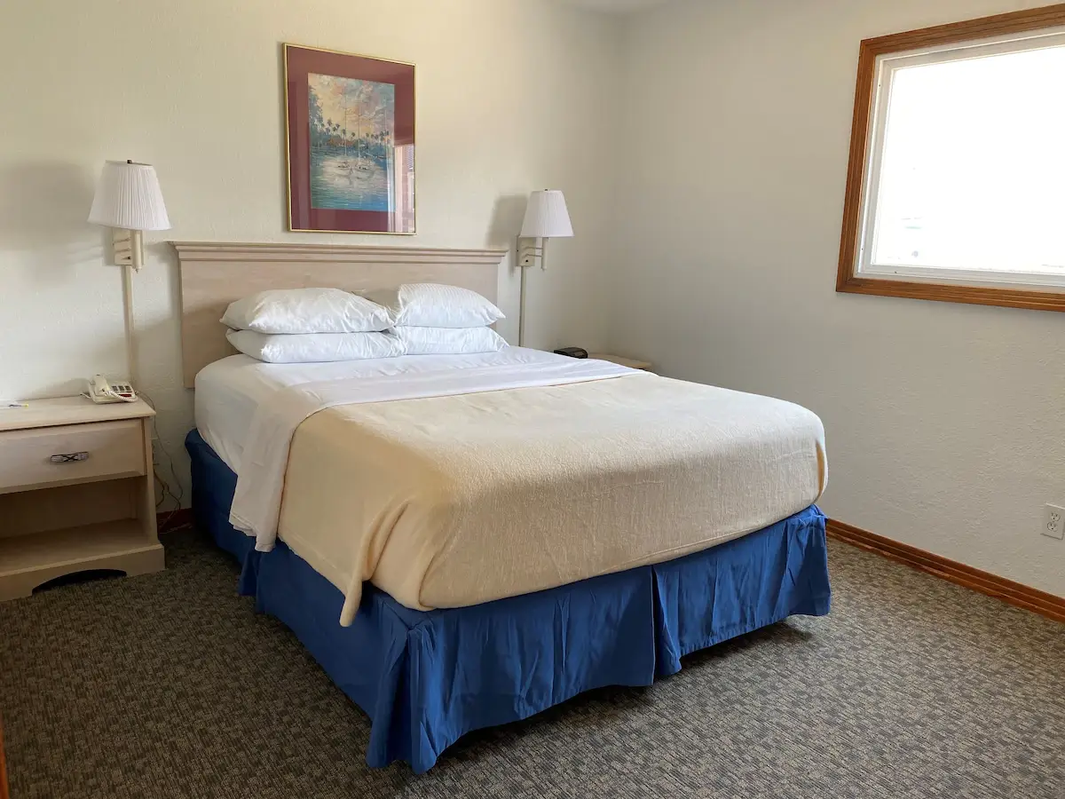 A bed with a cream coverlet and blue skirt at Mariner Inn & Suites, with a framed print of sailboats in a harbour above the headboard