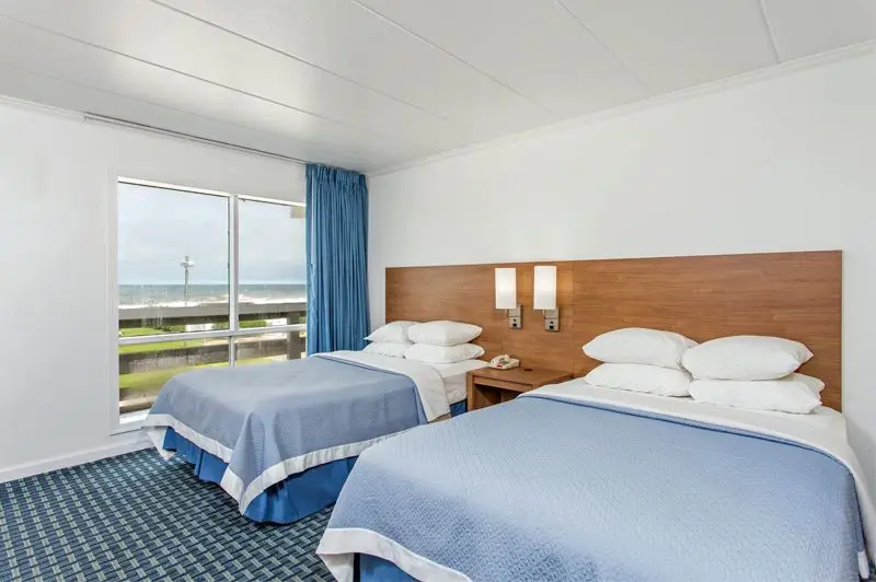 Two beds with blue coverlets in a room at Mariner Inn & Suites, beside a window with a blue curtain looking toward the ocean