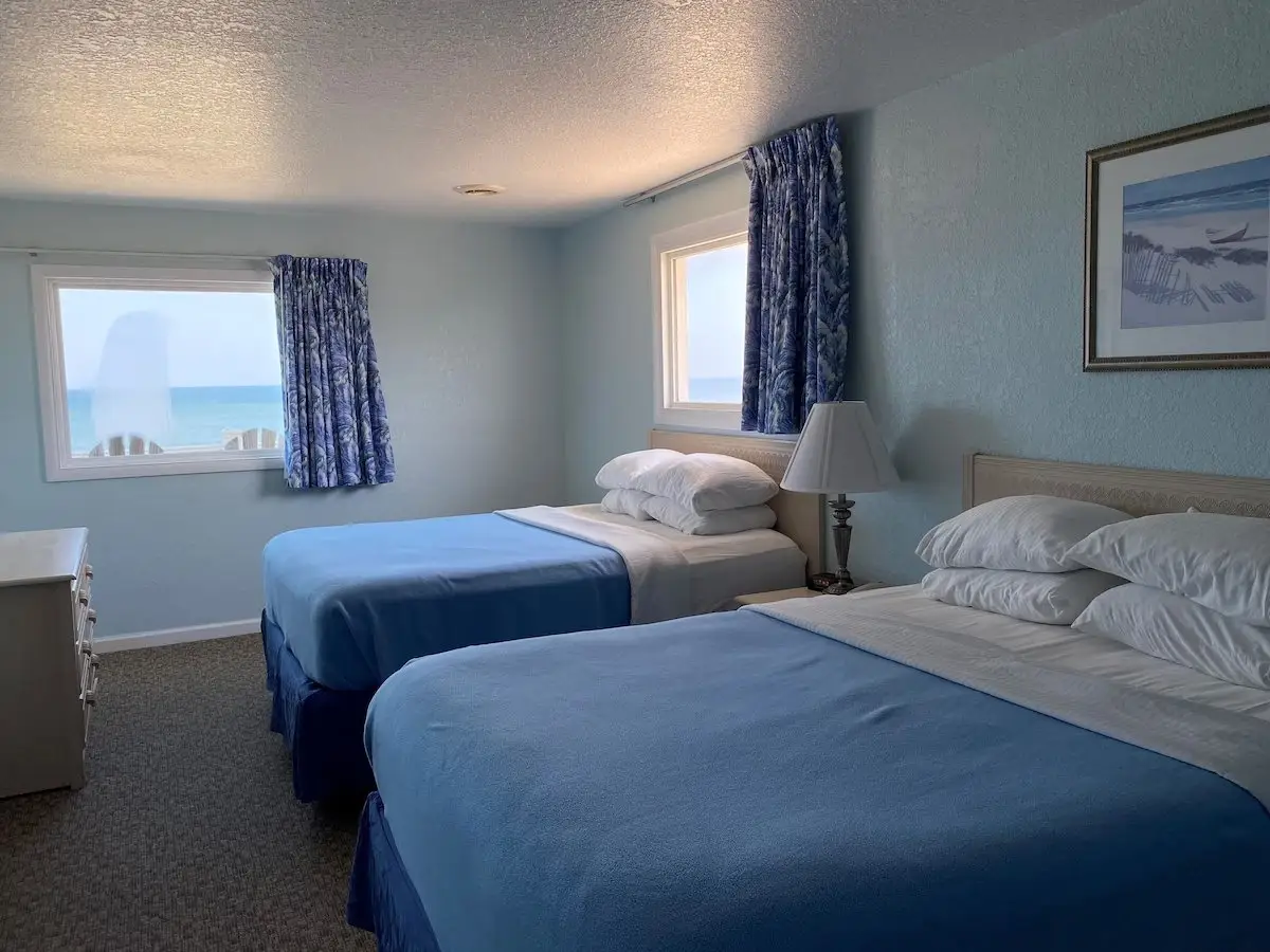 Two beds with steel-blue coverlets at Mariner Inn & Suites, with a framed beach photograph above the headboard and an ocean-view window with a palm-print curtain