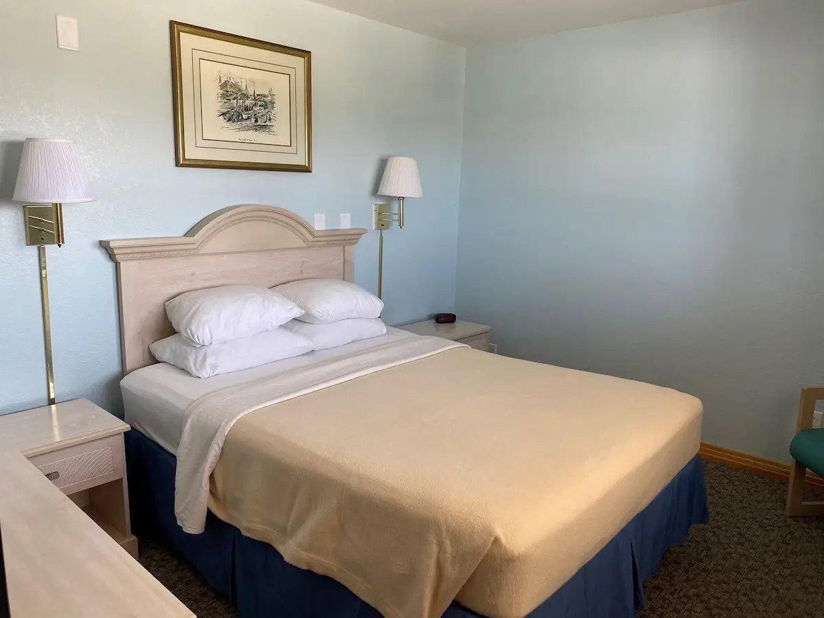 A bed with a pale gold coverlet and navy skirt at Mariner Inn & Suites, with a black-and-white dockside sketch above the headboard