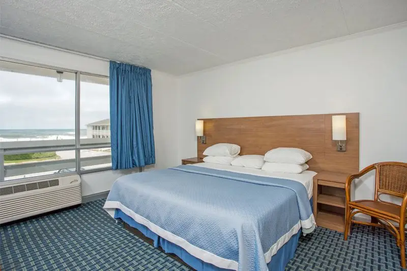 A king bed with a blue coverlet at Mariner Inn & Suites beside a window with a blue curtain, a rattan chair and a wall-mounted air conditioner