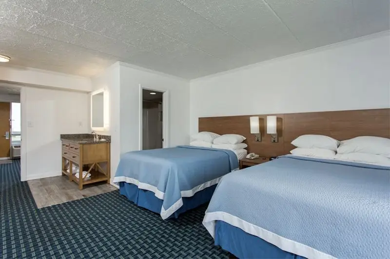 Two beds with blue coverlets in a room at Mariner Inn & Suites, with a wood vanity and mirror nook to the left