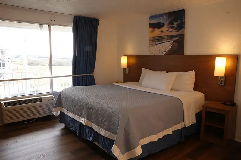 A king bed with a grey coverlet at Mariner Inn & Suites, with a sunset beach canvas above the headboard and a private balcony visible through the sliding glass door