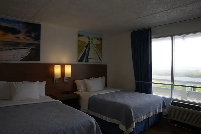 Two beds with grey and blue coverlets at Mariner Inn & Suites, with sunset-beach and dune-boardwalk canvas prints above the headboards