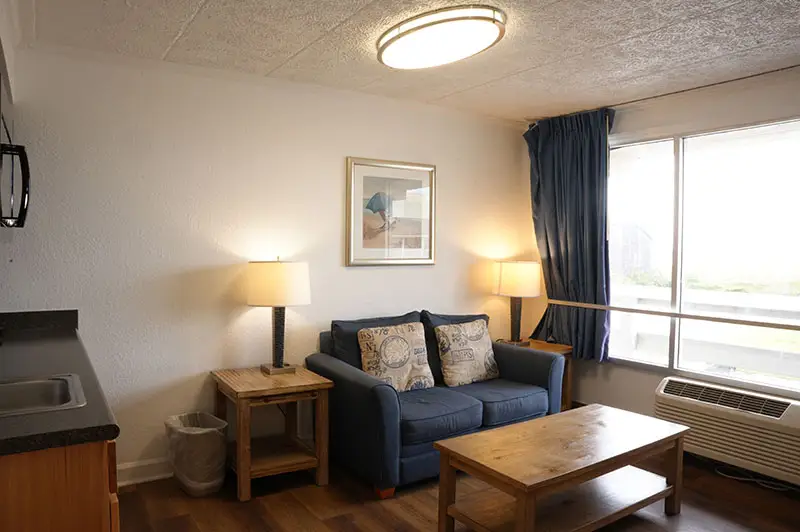 A suite living room at Mariner Inn & Suites with a navy loveseat, a beach-chair canvas print, and an ocean-view window with a navy curtain
