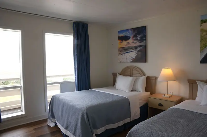 Two beds with grey and blue coverlets at Mariner Inn & Suites, with a sunset-beach canvas print and a private balcony visible through the window