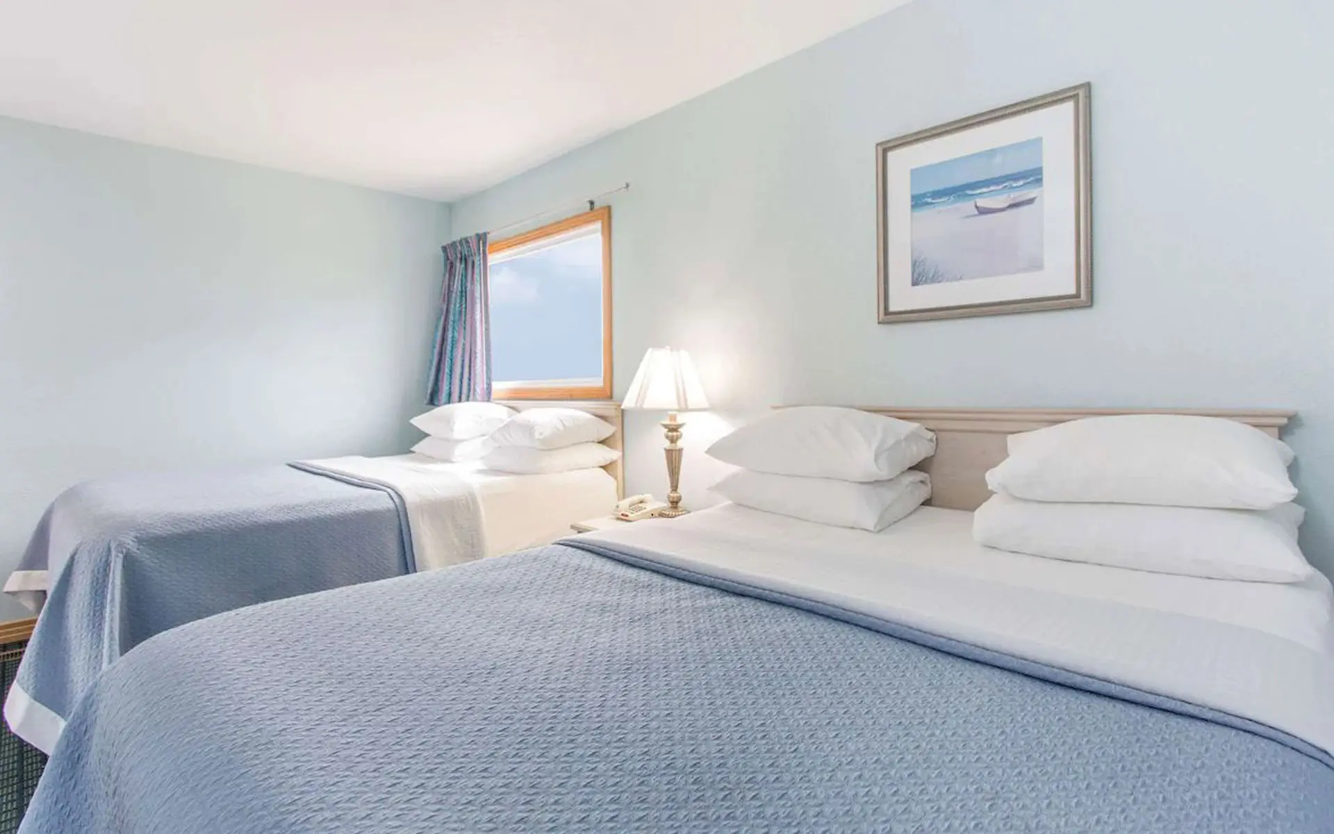 Two beds with blue coverlets in a guest room at Mariner Inn & Suites, with a framed beach print on the wall